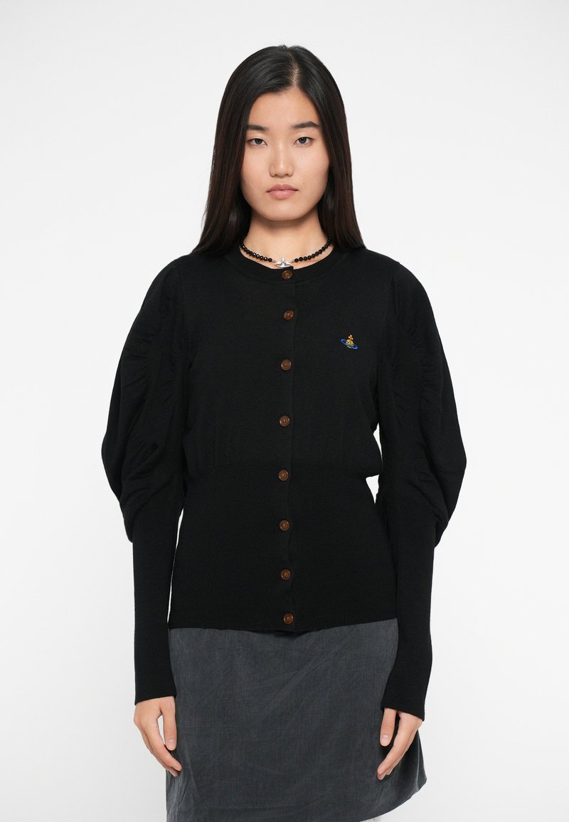 Cardigan nero a costine con maniche a sbuffo, dotato di bottoni marroni e un piccolo design ricamato. Indossato sopra una gonna grigia.