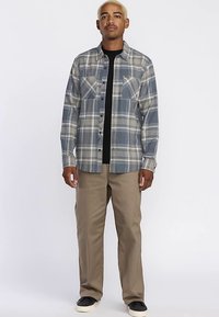 Chemise en flanelle à carreaux dans des tons bleus et gris, avec deux poches poitrine et une fermeture à boutons, associée à un pantalon beige et des chaussures noires.