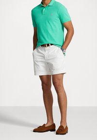 Mies, joka pitää päällään turkoosia polo-paitaa, valkoisia shortseja, ruskeaa vyötä, ruskeita tupsuläpsyjä ja kelloa sinisellä hihnalla, seisoo kädet taskuissa.