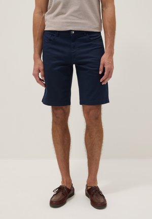 Hombre con pantalones cortos azul marino, camiseta beige y zapatos de cuero marrón con cordones, de pie frente a un fondo claro y liso.