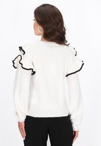 Witte trui met ruches aan de mouwen afgewerkt met zwarte trim. Heeft een ontspannen pasvorm en ribgebreide manchetten, met een zachte textuur en geen zichtbare patronen.