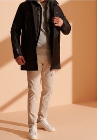 Parka acolchada negra con cremalleras, suéter gris, pantalones beige y zapatillas blancas. Textura suave, corte relajado y diseño en capas.
