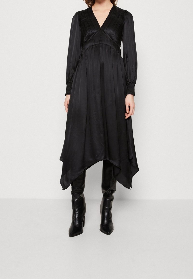Femme portant une longue robe noire avec un col en V, des manches longues, un ourlet en pointe et des bottes noires montantes à talons.