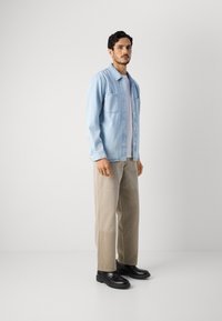 Lyseblå denimskjorte, med knapper og to brystlommer, båret over en grå t-shirt. Beige vide bukser og sorte sko.