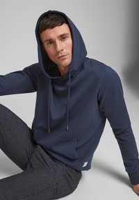 Haut à capuche bleu marine avec cordons et poche avant, associé à un pantalon à carreaux gris texturé. Tissu doux, coupe décontractée, branding minimal.