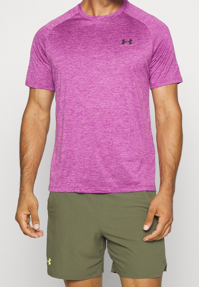 Muž v melírovaném růžovém sportovním tričku s krátkým rukávem a olivově zelených šortkách s logy na levé části hrudi a levé stehně.