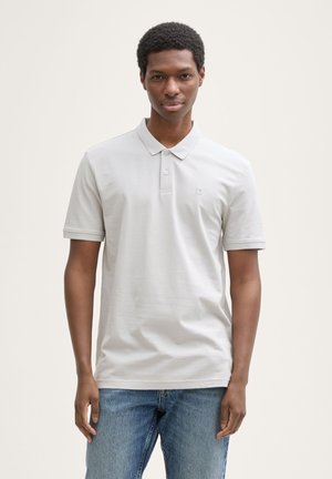 Hellgraues Poloshirt mit Knopfleiste, kurzen Ärmeln und einem dezenten Logo auf der Brust. Kombiniert mit blauen Jeans. Weiche Textur.