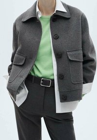 Grå uldblandingsjakke med store knapper, to forlommer, lagd over en grøn sweater og hvid skjorte, parret med mørke bukser.