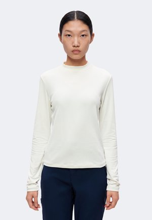 Jeune femme aux cheveux noirs et raides porte un haut blanc à manches longues uni et un pantalon sombre, debout devant un fond clair uni.