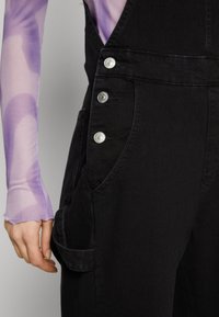 Black denim overalls feature silver snap buttons on the side and a side pocket. The fabric has a smooth texture with a fitted design.
