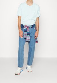 Světle modré bavlněné tričko s malým logem, v kombinaci se světle modrými džíny. Barevný svetr se zigzag vzorem uvázaný kolem pasu. Bílé tenisky.