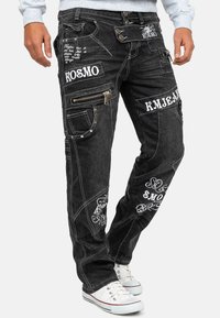 Jeans in denim nero con vari disegni ricamati, tasche con cerniera e cuciture decorative. Indossati con sneaker bianche.