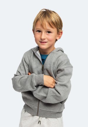Jeune garçon aux cheveux blonds portant un sweat à capuche gris zippé et un pantalon clair, les bras croisés, regardant légèrement en souriant vers la caméra.