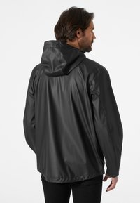 Chaqueta impermeable negra con capucha, que presenta una textura suave y un corte holgado. El diseño incluye costuras visibles y puños elásticos.