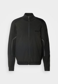 Black jacket with a stand collar, zip closure, textured fabric accents, side pockets, and logo on the chest. Features mesh details on sleeves.