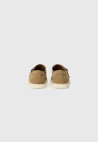 Chaussures en daim marron avec une profil bas, dotées d'une semelle en caoutchouc blanche et de coutures discrètes. Deux boucles décoratives sur le côté.