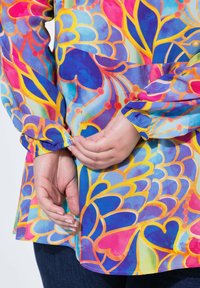 Blusa multicolore con motivi floreali astratti in blu, rosa, giallo e verde. Maniche elastiche e dettaglio a orlo svasato.