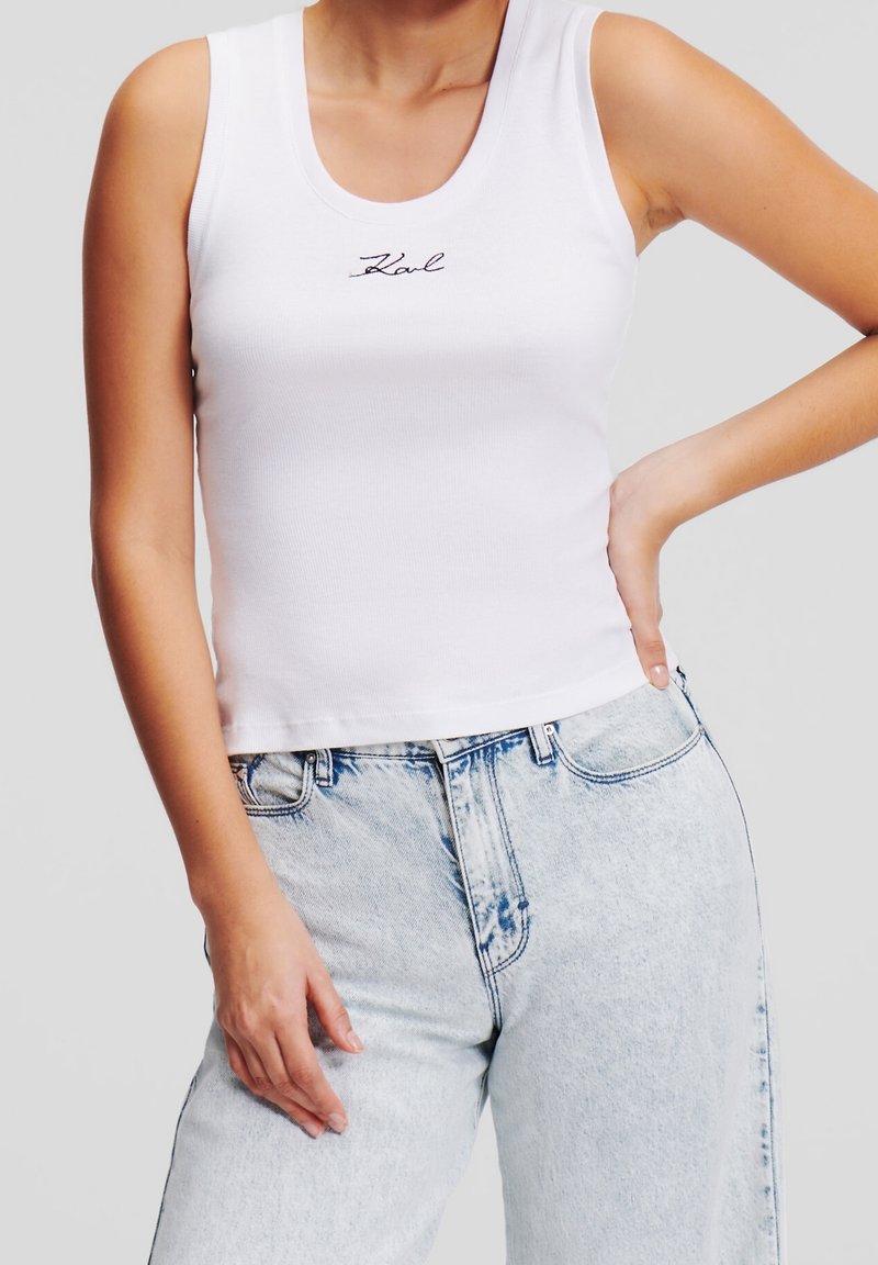 Femme portant un débardeur blanc sans manches avec "Karl" brodé et un jean taille haute bleu clair, main sur la hanche.