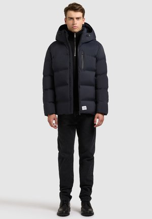 Marineblauwe pufferjack met een capuchon, voorzien van een voorrits, een borstzak en zij-elastische manchetten; gedragen over een zwart topje en jeans.