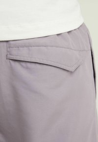 Gros plan sur la poche d'un pantalon violet clair avec un rabat incliné et des coutures détaillées, porté avec une chemise blanche rentrée.
