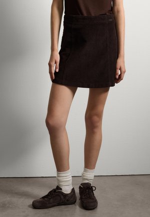 Mini falda de pana marrón con un diseño ajustado y detalles de costura en la parte frontal, combinada con zapatillas deportivas oscuras y calcetines blancos hasta el tobillo.