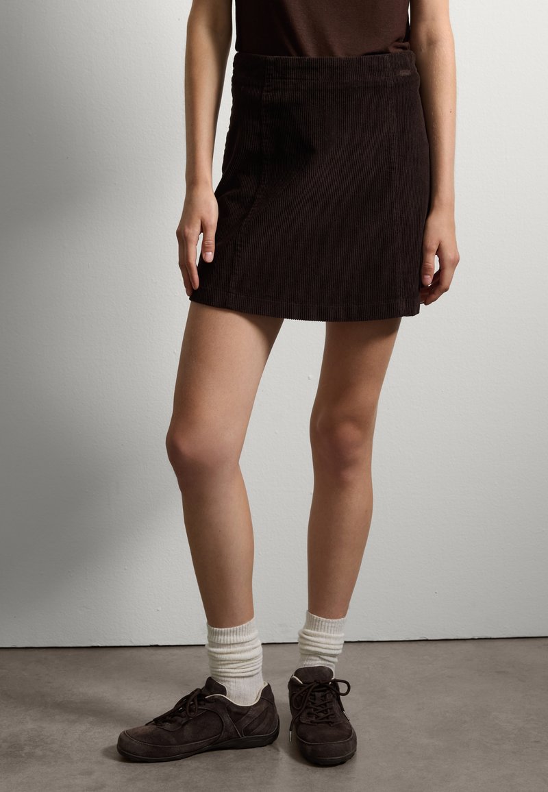 Mini falda de pana marrón con un diseño ajustado y detalles de costura en la parte frontal, combinada con zapatillas deportivas oscuras y calcetines blancos hasta el tobillo.
