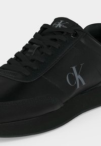 Baskets en cuir noir avec lacets noirs, avec le logo Calvin Klein "cK" sur le côté et la languette, sur un fond clair uni.