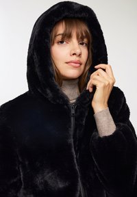 Giacca nera in pelliccia sintetica con cappuccio, dotata di zip sul davanti, caratterizzata da una texture morbida e maniche larghe, con un dolcevita neutro sottostante.