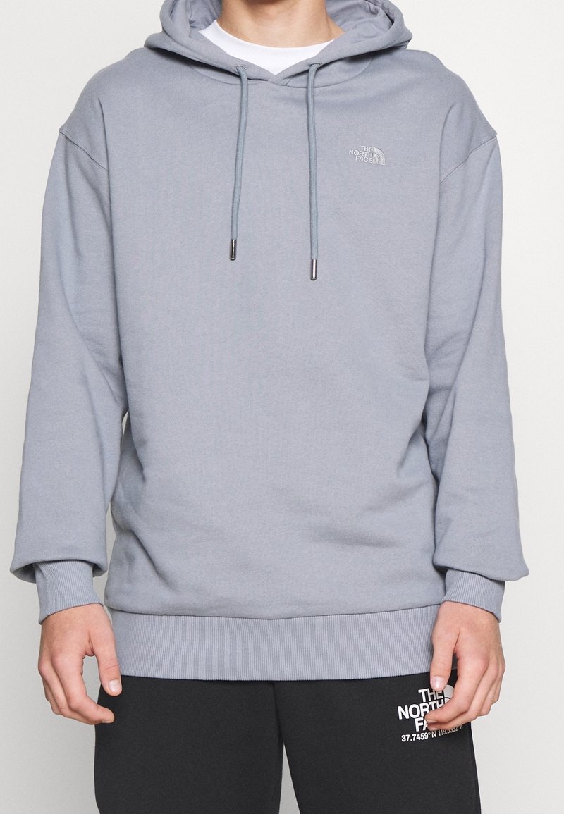 Sweat-shirt à capuche gris avec une coupe ample, poignets et ourlet côtelés, arborant un logo et des cordons de serrage avec des embouts métalliques. Matière douce en mélange de coton.