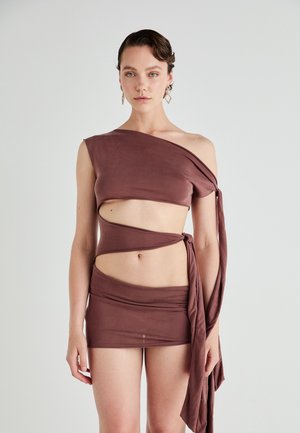 Braunes Zweiteiler-Outfit mit einem einseitigen Oberteil, das Ausschnitte und einen gebundenen Träger aufweist, kombiniert mit einem kurzen, figurbetonten Rock. Weiche Stofftextur.
