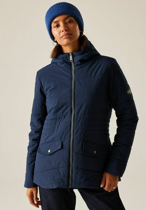 Kobieta ubrana w granatową pikowaną kurtkę na zamek z kieszeniami i niebieską dzianinową czapkę, stojąca na tle gładkiego jasnego tła.