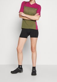 Tričko s krátkým rukávem má zelený a růžový barevný blok a velký nápis "MONS ROYALE". Kombinováno s černými kraťasy a cyklistickými botami.