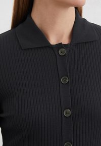 Schwarzes, geripptes Strickoberteil mit einem Kragen und fünf schwarzen Knopfverschlüssen auf der Vorderseite.