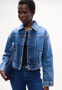 Jacket di jeans tagliato in blu sbiadito con bottoni argentati, due tasche frontali e colletto classico. Abbinato a un top scuro e una borsa blu.