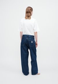 Camiseta blanca de manga corta combinada con jeans de mezclilla anchos de color azul. Los jeans cuentan con dos bolsillos traseros y un pequeño parche con el logo de la marca.