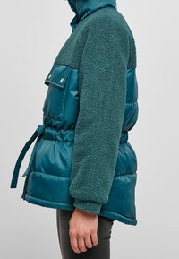 Tyrkysová péřová bunda s huňatým zeleným beránkem na vršku, se staženým pasem na šňůrku a dvěma předními kapsami se zlatými kovovými doplňky.