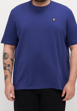 Mann trägt ein schlichtes blaues Kurzarm-T-Shirt mit einem kleinen gelben Vogel-Logo auf der Brust und sichtbaren Tätowierungen am linken Unterarm.