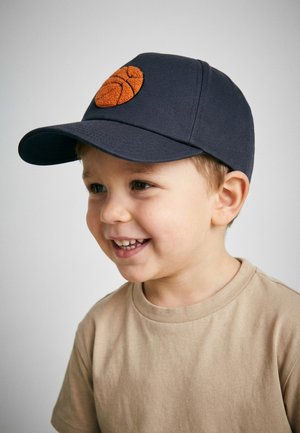 Smiling young boy wearing a beige t-shirt and navy blue cap with an orange basketball patch, looking slightly to the right.