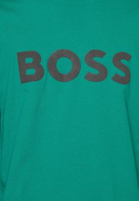 Teal bomuld t-shirt med store sorte bogstaver, der stavner "BOSS" hen over forsiden. Kort ærme og standard rund halsudskæring.