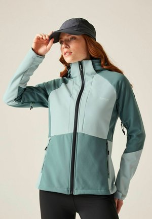 Kobieta w dwukolorowej zielonej kurtce outdoorowej i czarnej czapce, poprawiająca czapkę jedną ręką, stojąca na jednolitym tle.