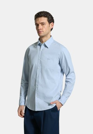 Mann trägt hellblaues Hemd mit Knöpfen und dunkelblaue Hose, steht mit einer Hand in der Tasche vor einem schlichten hellen Hintergrund.