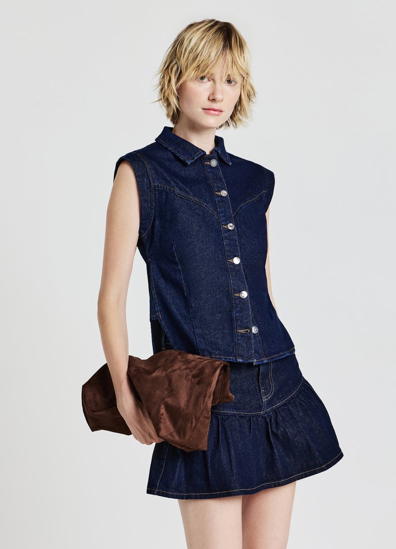 Top senza maniche in denim scuro con chiusura a bottoni, abbinato a una gonna in denim con volant. Il modello tiene un accessorio marrone con una texture morbida.