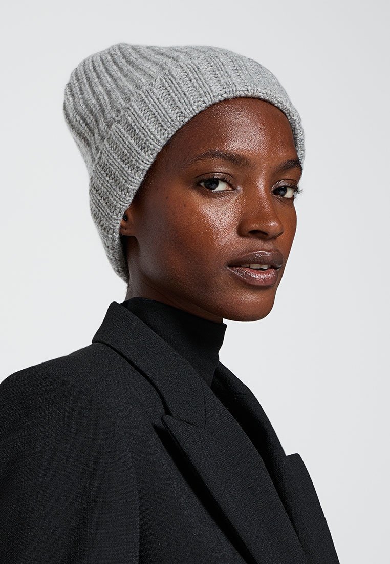 Jasnoszara, dziana czapka beanie o prążkowanej strukturze, lekko opadający fason, zestawiona z czarnym żakietem i bluzką z wysokim kołnierzem. Czyste tło.