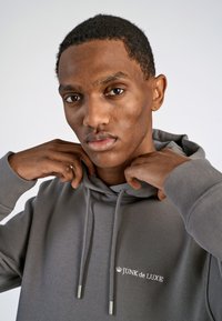 Grijze hoodie met verstelbare trekkoorden en het logo "JUNK de LUXE" in het wit. Heeft een zachte textuur en ribbelmanchetten.