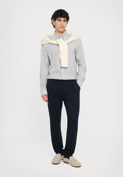 Chemise à rayures bleu et blanc, pull crème drapé autour du cou, pantalon marine et mocassins beiges.