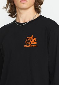 Schwarzes Langarmshirt mit einem auffälligen orangen Grafikdesign, das einen Stern und das Wort "Workwear" auf der Brust zeigt. Weicher Baumwollstoff.