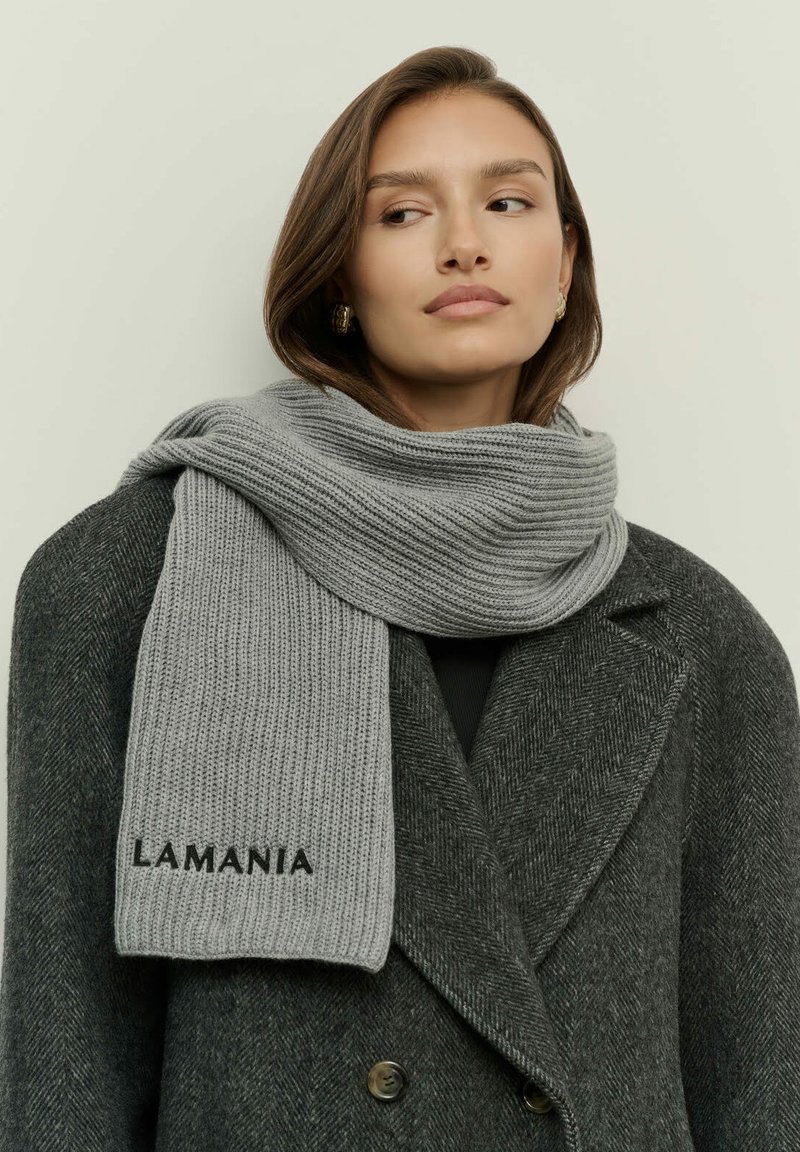 Egy nő sötét szürke halszagú kabátot és világosszürke kötött sálat visel, amin a "LAMANIA" felirat látható, oldalra nézve egy sima háttér előtt.