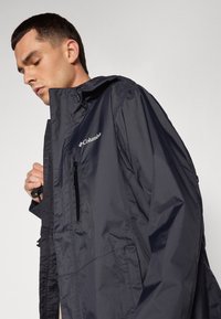 Veste imperméable marine avec une poche poitrine zippée, une capuche réglable et des poignets élastiques. Présente un logo sur la partie gauche de la poitrine.