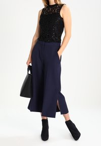 Haut sans manches noir à sequins, pantalon large bleu marine avec fentes, et bottines noires. Porte un sac à main noir.