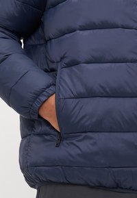 Asmens ranka užtraukiamoje kišenėje tamsiai mėlyne siuvinėtos pūkinės striukės su elastinėmis rankovėmis ir juosmeniu.